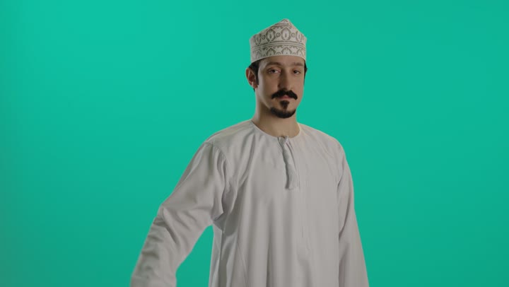 الوقوف بثقة مع ثني الذراع , النظر لعضلات الذراع دليل على القوة الجسدية , رجل عربي خليجي عماني يرتدي دشداشة بيضاء و كمة مطرزة , رفع الحاجب لاعلى و نظرة الثبات و الثقة