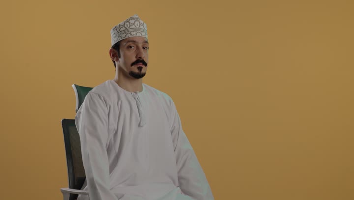 النظر للكاميرا مع تعابير مبتسمة راضية , رجل عربي خليجي عماني يرتدي دشداشة بيضاء و كمة مطرزة يرفع اصبع الابهام لاعلى علامة على الاعجاب بشئ ما , ايماءات سعادة و بهجة واضحة