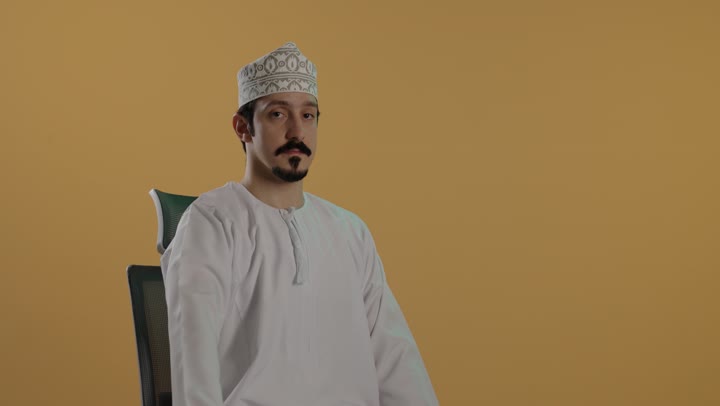 رفع الذراع و توجيه اصبع الابهام للاسفل اشارة الى عدم الاعجاب بشئ ما , رجل عربي خليجي عماني يرتدي كمة مطرزة و الدشداشة البيضاء يجلس على الكرسي , تعابير وجه محبطة و غير راضية