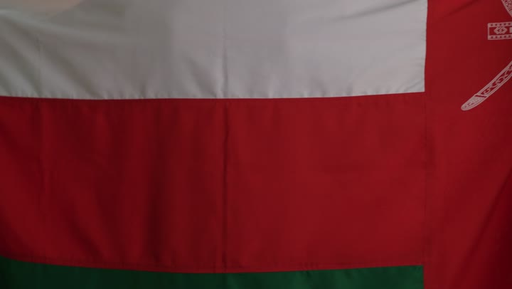 شاب عربي خليجي عماني يرتدي دشداشة بيضاء و كمة مطرزة يرفع العلم العماني على الاكتاف اشارة الة حب الوطن , الالتفاف للكاميرا , الوقوف بشموخ  , تعابير العزة و الفخر