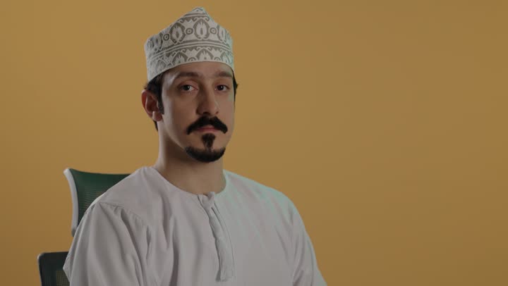 رفع الذراع للاعلى مع فتح الكف  امام الكاميرا اشارة على التوقف و الامتناع عن القيام بشئ ما , رجل عربي خليجي عماني يرتدي دشداشة بيضاء وكمة مطرزة تظهر علو وجهه تعابير عدم الرضا و الاستياء