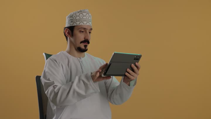 Expressions of happiness and a smile after completing the required tasks, an Arab Gulf Omani man wearing a white dishdasha and an embroidered kumma, holding a sophisticated tablet to perform the task easily while sitting in the office.