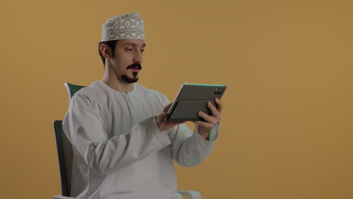 الدقة و التركيز اثناء القيام بالمهام المطلوبة , رجل عربي خليجي عماني يرتدي الدشداشة البيضاء و كمة مطرزة يحمل الجهاز اللوحي المتطور , تعابير الجدية و الاحترافية