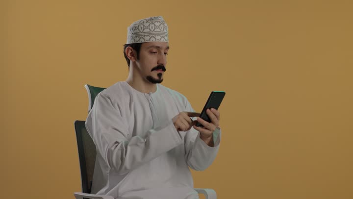 استخدام التطبيقات المختلفة للقيام بالاعمال المتعددة باستخدام جهاز تقني واحد , رجل عربي خليجي عماني يرتدي الدشداشة البيضاء و الكمة المطرزة يحمل الهاتف المحمول و يقوم باجراء المكالمات الجماعية , تعابير السعادة ظاهرة على الوجه