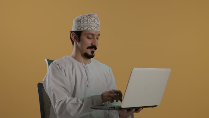 أداء الاعمال المكتبية بدقة باستخدام الاجهزة المتطورة , رجل عربي خليجي عماني يرتدي دشداشة بيضاء و كمة مطرزة يحمل الحاسب المتنقل بخفة , تعابير الابتسام و الجدية