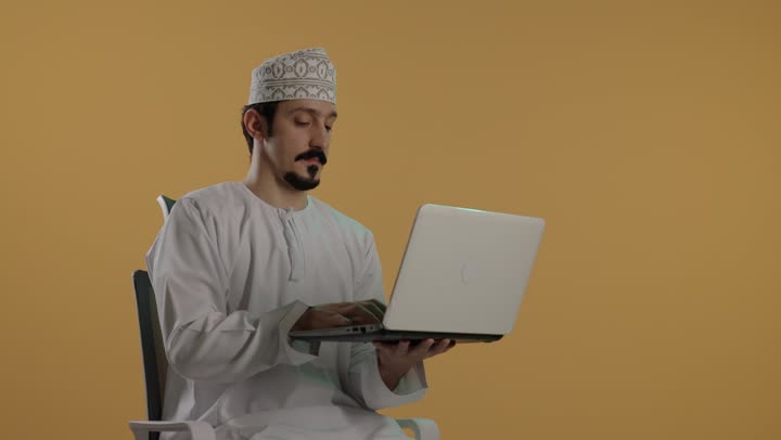 تعابير السعادة و الابتسام للكاميرا اثناء القيام بالاعمال المكتبية بسهولة , شاب عربي خليجي عماني يرتدي الدشداشة البيضاء و الكمة المطرزة يحمل الحاسوب المتنقل بخفة , ايماءات البهجة باستخدام الاجهزة التقنية المتطورة