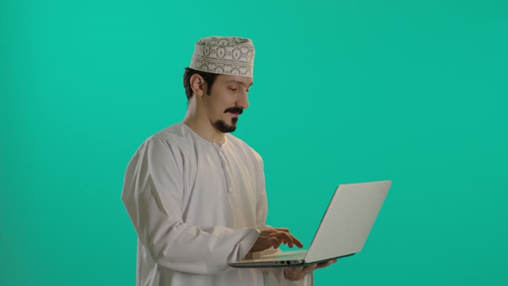Writing performance tasks with professionalism and precision, an Arab Gulf Omani man wearing a white dishdasha and an embroidered kumma holds the technical device with ease and lightness, with a happy facial expression, looking at the camera.