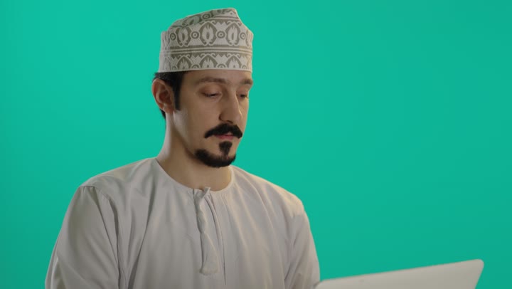 التركيز و الدقة في استخدام الاجهزة التقنية الحديثة , رجل عربي خليجي عماني يرتدي الدشداشة البيضاء و كمة مطرزة يحمل الحاسوب المتنقل بسهولة ,