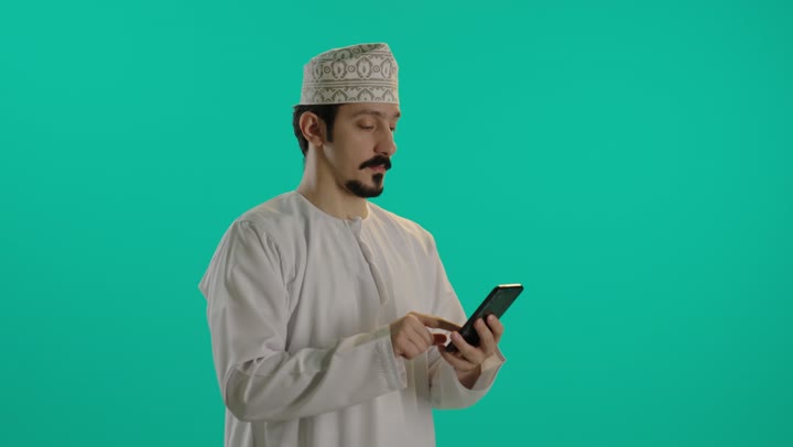 البقاء متصلاً مع العالم الخارجي الكبير و معرفة الاخبار العالمية بسهولة , رجل عربي خليجي عماني يرتدي الدشداشة البيضاء و الكمة المطرزة  يتصفح الهاتف المحمول ,  ايماءات السعادة و السرور , النظر للكاميرا و الابتسامة