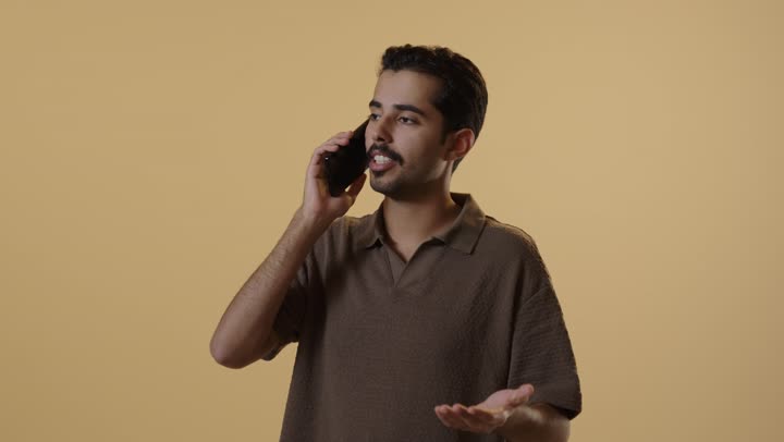 Speaking with enthusiasm and passion, a Gulf Arab Saudi man dressed in casual attire is making a phone call, with expressions of joy and happiness while moving his hand as a sign of feeling excited.