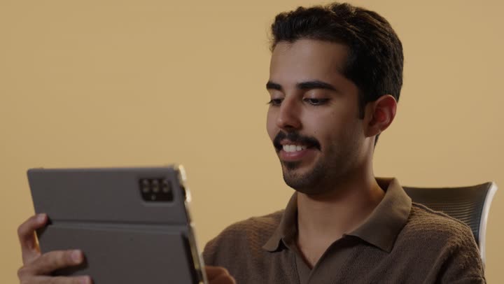 تعابير سرور و بهجة ظاهرة على وجه الشاب , رجل عربي خليجي سعودي يرتدي الزي الكاجوال يستخدم الجهاز اللوحي لاغراض متعددة و مفيدة