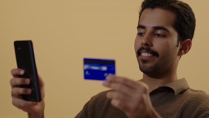 Entering the secret numbers of the credit card to purchase products online, a Gulf Arab Saudi man dressed in casual attire is starting the online shopping process, showing gestures of excitement and happiness.