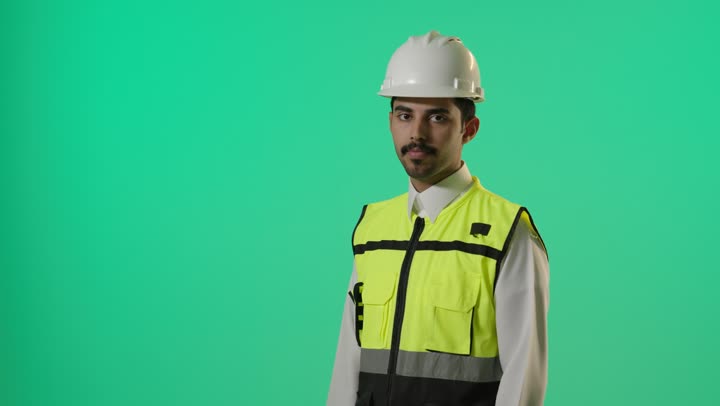 النظر الى الكاميرا مع الوقوف بثبات وثقة , مهندس عربي خليجي سعودي يرتدي اليونيفورم خاص بالمهندسين يعقد ذراعيه , تعابير الثبات و الاصرار
