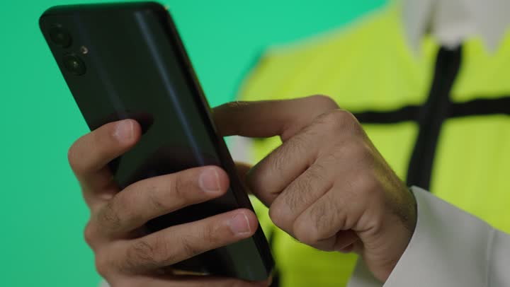 التصفح على الهاتف المحمول أثناء العمل لمعرفة اخر المستجدات في عالم الهندسة , مهندس عربي خليجي سعودي يرتدي الثوب الابيض و الصديري العاكس للضوء يتصفح الجوال الاسود , تصوير مقرب