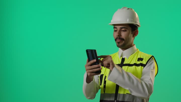 Conducting group video calls among team members to accomplish the task, an Arab Gulf Saudi engineer wearing a white thobe and safety helmet at work sites, using various applications to stay connected, expressions of happiness and joy.