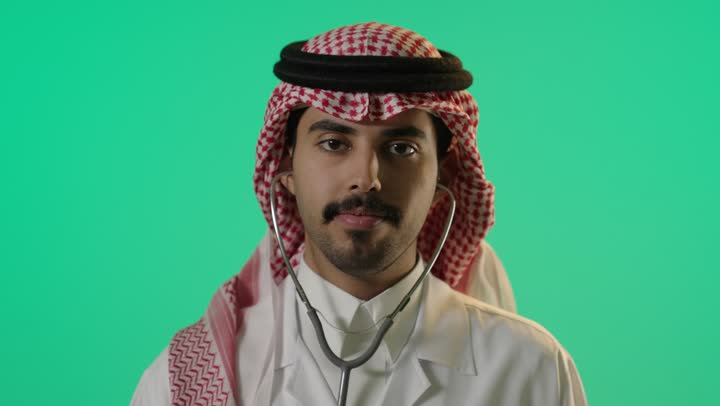 طبيب عربي خليجي سعودي يرتدي المعطف الابيض الخاص بالاطباء , وضع السماعة الطبية في الاذن والتوجيه بها للكاميرا  , تعابير السرور و البهجة أثناء مزاولة مهنة الطب