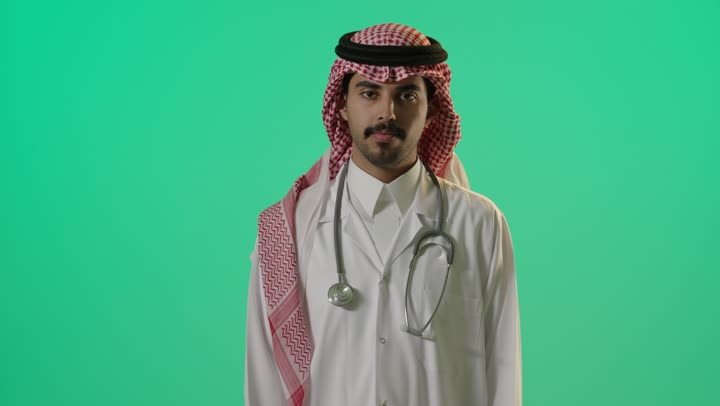 Raising the thumb up is a sign of liking something, an Arab Gulf Saudi doctor wearing a white medical coat and placing the stethoscope on his shoulders, expressions of happiness and joy.