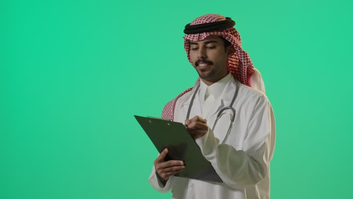 طبيب عربي خليجي سعودي يرتدي الثوب الابيض التقليدي في المملكة العربية السعودية , كتابة التقارير الطبية بتركيز و حرفية , ايماءات السعادة اثناء قضاء ساعات الدوام اليومي