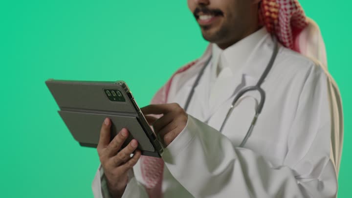 Communication with doctors all over the world, an Arab Gulf Saudi doctor wearing a white medical coat and traditional attire in the kingdom browsing the latest in the field of medicine, using a tablet to expand the circle of knowledge, expressions of joy and passion for learning.