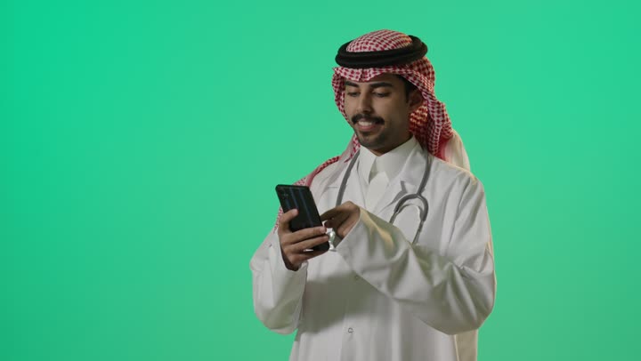 استخدام الاجهزة التقنية الحديثة لقضاء وقت مسلي في أوقات الراحة , طبيب عربي خليجي سعودي يرتدي المعطف الطبي الابيض و الثوب التقليدي في المملكة يحمل الهاتف المحمول في المحادثات الجماعية و الاتصالات , التواصل مع العائلة و الاصدقاء , تعابير السرور و الابتهاج