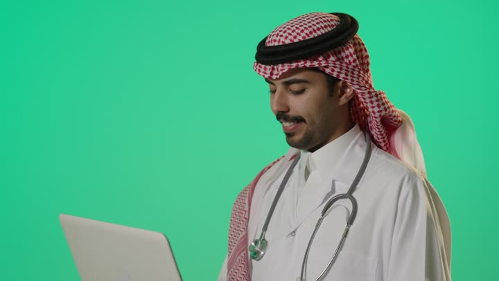 سرعة الوصول الي المعلومات الجديدة و ارسال البريد الالكتروني الي باقي فريق العمل , طبيب عربي خليجي سعودي يرتدي المعطف الطبي الابيض و الثوب التقليدي في المملكة يحمل الحاسب المتنقل , تعابير وجه سعيدة و فرحة , الاحترافية و الاتقان في استخدام الاجهزة التقنية الحديثة