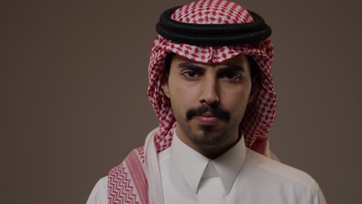 تعابير وجه غاضبة مستاءة , رجل عربي خليجي سعودي يرتدي الثوب التقليدي الابيض يقف امام الكاميرا , رفع اصبع السبابة امام الفم علامة على طلب السكوت و التوقف عن الكلام , ايماءات متجهمة مع عقد الحاجبين