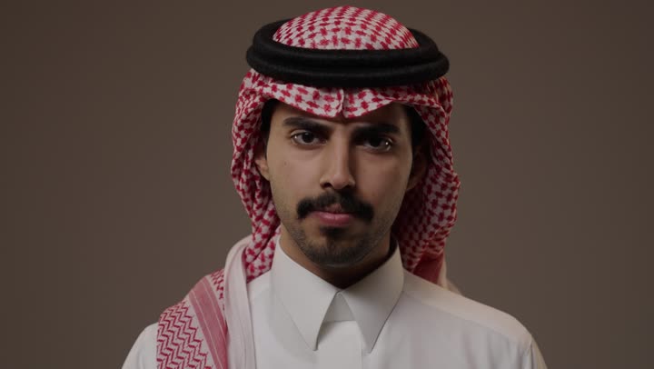 وضع اصبع السبابة امام الفم اشارة الي طلب التوقف عن الكلام, رجل عربي خليجي سعودي يرتدي الزي التقليدي الابيض للمملكة العربية السعودية , الوقوف امام الكاميرا , تعابير وجه غاضب متجهم