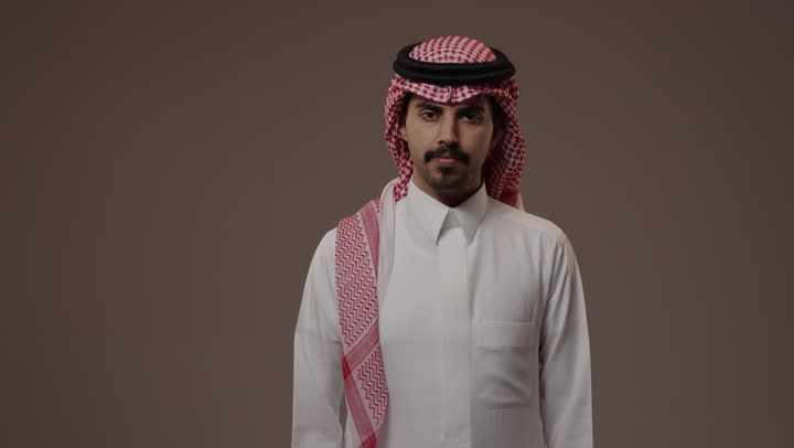 استعراض القوة الجسدية اثناء الوقوف امام الكاميرا , رجل عربي خليجي سعودي يرتدي الثوب الابيض التقليدي في المملكة العربية السعودية يثني ذراعه و يظهر عضلاته , تعابير القوة و الثبات