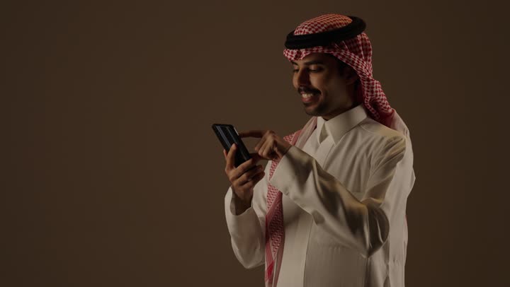 Checking on family and friends through various communication and chat applications, an Arab Saudi Gulf man wearing the traditional white Saudi thobe and a colored ghutrah holds a mobile phone, with expressions of happiness and joy, spending enjoyable times using different technological devices.