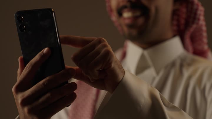 الشعور بالسعادة و الفرحة اثناء ارسال الرسائل النصية و المحادثات , رجل عربي خليجي سعودي يرتدي الزي التقليدي الابيض يحمل الهاتف المحمول  , تصوير مقرب , تعابير الابتسامة و الابتهاج