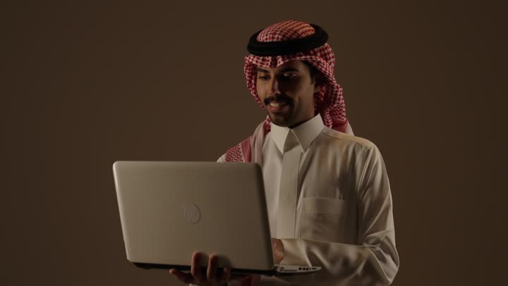 استخدام الاجهزة المتطورة في الدوام اليومي , رجل عربي خليجي سعودي يرتدي الثوب الابيض التقليدي في المملكة يحمل الحاسوب المتنقل بسهولة و خفة , ارسال الرسائل و البريد الالكتروني  , تعابير السرور و الفرحة