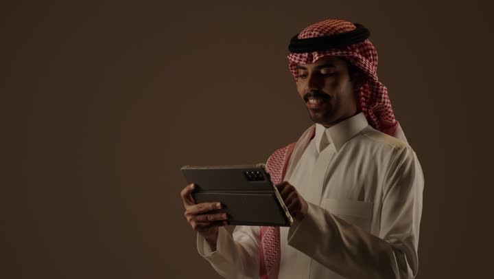 رجل عربي خليجي سعودي يرتدي الثوب الابيض التقليدي في المملكة العربية السعودية يحمل الجهاز اللوحي الخفيف بسهولة , متابعة المستجدات اول بأول عبر ارسال الرسائل النصية و استخدام التطبيقات الخاصة بها , تعابير السعادة و البهجة , وجه مبتسم سعيد