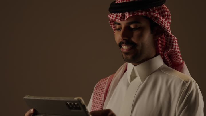 البقاء متصل بالعائلة و الاصدقاء باستخدام التطبيقات المتعددة الخاصة بارسال الرسائل النصية و معرفة الاخبار  , رجل عربي خليجي سعودي يرتدي الزي التقليدي في المملكة يحمل الجهاز اللوحي و يتابع اخر المستجدات , ايماءات السرور و الفرحة