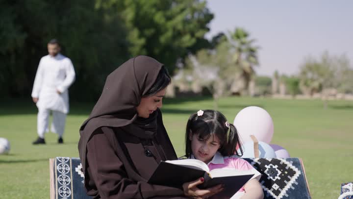تقضية اوقات ممتعة تحت السماء الصافية و الشمس الساطعة , رجل عربي خليجي سعودي يرتدي الثوب التقليدي الابيض يلعب كرة القدم مع ابنه في مرح و سعادة , امرأة عربية خليجية سعودية محجبة ترتدي عباءة سوداء تقرأ مع ابنتها القصص المسلية , مفهوم الوقت القيم بين افراد العائلة