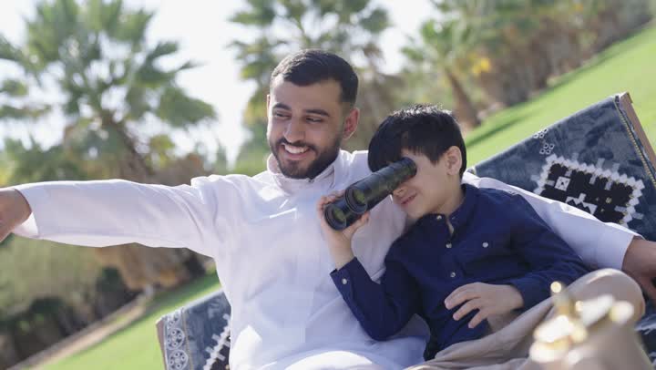 اب عربي خليجي سعودي يرتدي ثوب تقليدي ابيض يشير الى الاشجار البعيدة , ولد عربي سعودي خليجي يرتدي الزي الكاجوال يمسك المنظار لرؤية شئ بعيد , محادثة ودية بين الاب و ابنه تحت السماء الصافية