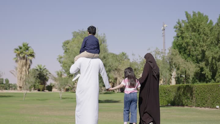 الاب القائد يحمل ابنه على الاكتاف و يمسك بيد باقي افراد العائلة , امرأة عربية خليجية سعودية محجبة ترتدي عباءة سوداء تمسك يد ابنتها الصغيرة , التنزه بين المساحات الخضراء الواسعة , رجل عربي خليجي سعودي يرتدي ثوب ابيض تقليدي يقود المسيرة وسط الطبيعة الخلابة و الاجواء الدافئة