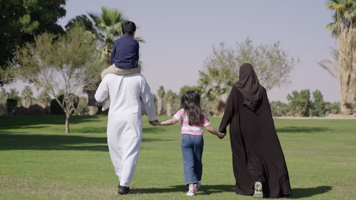 عائلة عربية خليجية سعودية تمسك بيد بعضها اشارة على الترابط الاسري , تعدد مصادر الخروجات العائلية في المملكة , رجل عربي سعودي خليجي يرتدي ثوب تقليدي ابيض يحمل ابنه على الاكتاف , طفلة عربية خليجية سعودية ترتدي الزي الكاجوال تتمسك بيد الام و الاب  , التصوير من زاوية خلفية وسط المساحات الخضراء , التجول تحت الشمس الساطعة
