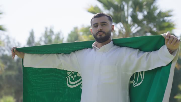 حمل العلم السعودي على الاكتاف اشارة الى عقد النية على رفع شأن الوطن عالياً , رجل عربي خليجي سعودي يرتدي ثوب عربي تقليدي يتوشح بالراية الخضراء السعودية , الاجواء المشمسة الدافئة ,