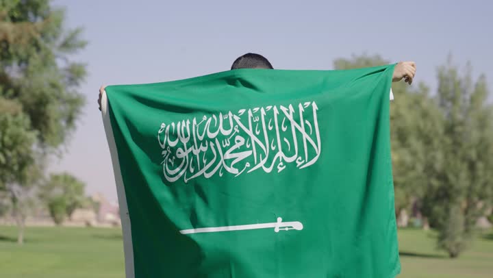 رجل عربي خليجي سعودي يرتدي ثوب تقليدي ابيض يلتفت للكاميرا و يبتسم اثناء حمل العلم السعودي على الاكتاف , الاحتضان براية التوحيد لتعزيز الشعور بالامان