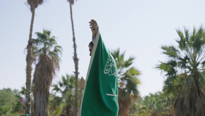 الالتفاف و النظر للكاميرا مع ابتسامة العزة اثناء حمل العلم السعودي , رجل عربي سعودي خليجي يرتدي الثوب التقليدي الابيض يحمل على ظهره راية التوحيد الخضراء