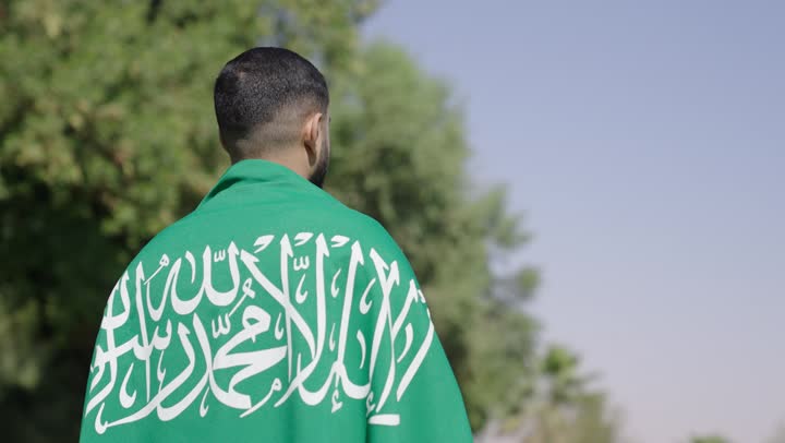 Enjoying the warm atmosphere inside safe homelands, an Arab Gulf Saudi man wearing a traditional white thobe carries the Saudi flag on his back as a sign of embrace and a sense of security.