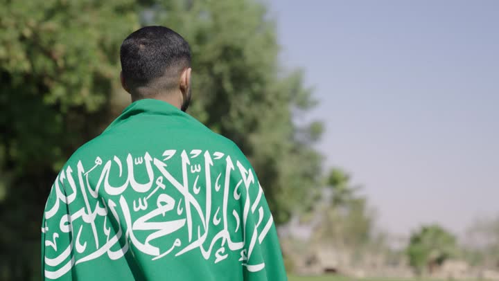 A Saudi Arabian Gulf man wearing a traditional white thobe feels proud while holding the Saudi flag. There are various family activities within the kingdom to spend special times.
