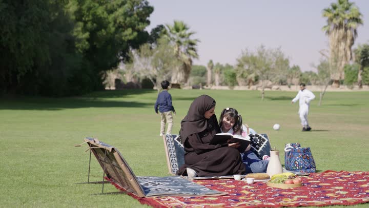 طلعات اسرية قيمة بين الطبيعة و الاشجار العالية , رجل عربي خليجي سعودي يرتدي الثوب التقليدي الابيض يلعب بالكرة مع ابنه بمرح و سعادة , سيدة عربية خليجية سعودية محجبة ترتدي عباءة سوداء تجلي بجوار ابنتها الصغيرة و تقرأ معها القصص المسلية , جو مشمس و دافئ , اوقات عائلية مميزة