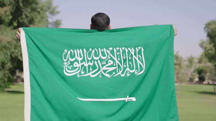 A rear angle shot of an Arab Gulf Saudi man wearing a traditional white thobe, carrying the Saudi flag on his back, in warm sunny weather, with a clear sky above a safe and secure homeland.