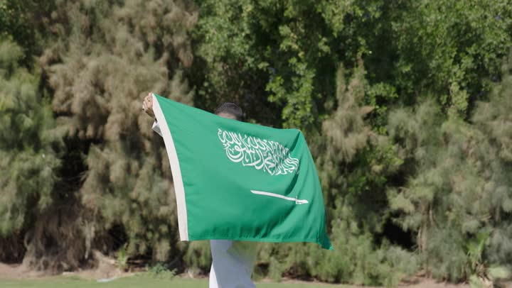 The green banner of monotheism flies high in the clear sky, a Saudi Arabian Gulf man wearing a traditional white thobe runs amidst the enchanting nature, carrying the Saudi flag over his shoulders, in a warm atmosphere that is pleasing to the soul.