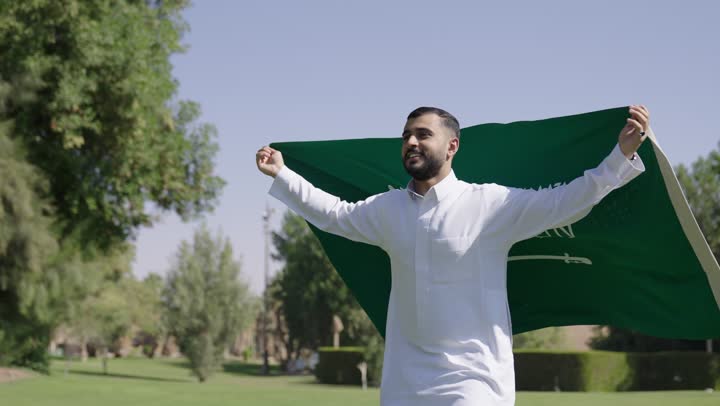 ايماءات السعادة و الفخر , رجل عربي خليجي سعودي يرتدي الثوب الابيض التقليدي يحمل العلم السعودي و يجري تحت الشمس الدافئة , الشعور بالامان و الطمأنينة داخل الوطن العزيز