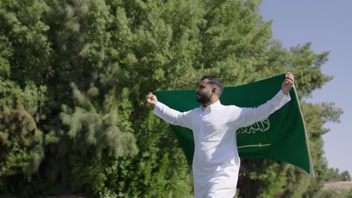 الشعور بالفخر و الاعتزاز اثناء رفع الراية التوحيد الخضراء , رجل عربي خليجي سعودي يحمل العلم السعودي على اكتافه و يجري وسط الحدائق الواسعة , الشعور بالامن و الامان في الوطن الحبيب