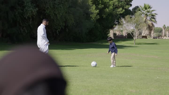 قراءة الكتب التاريخية لمعرفة معلومات قيمة عن تاريج المملكة , تعليم و تسلية , ام عربية خليجية سعودية محجبة ترتدي العباءة السوداء تجلس بجوار الابنة الصغيرة تقرأ لها القصص التاريخية التراثية عن الاجداد , اب عربي خليجي سعودي يرتدي الثوب الابيض التقليدي يركل  الكرة مع ابنه الصغير , الاستمتاع بالنزهة العائلية وسط الطبيعة الساحرة