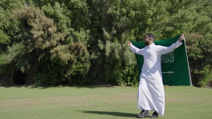 النظر الى الطبيعة الساحرة و الاستمتاع بالاجواء المحببة , رجل عربي خليجي سعودي يرتدي الثوب التقليدي الابيض يحمل العلم السعودي على اكتافه في فخر و عزة , الجري تحت السماء الصافية والشمس الساطعة