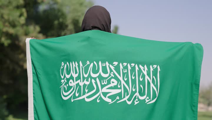 الدوران في حركات دائرية اثناء حمل العلم السعودي فوق الاكتاف , امراة عربية خليجية سعودية محجبة ترتدي عباءة سوداء تمسك راية التوحيد الخضراء و تدور بها وسط المساحات الخضراء